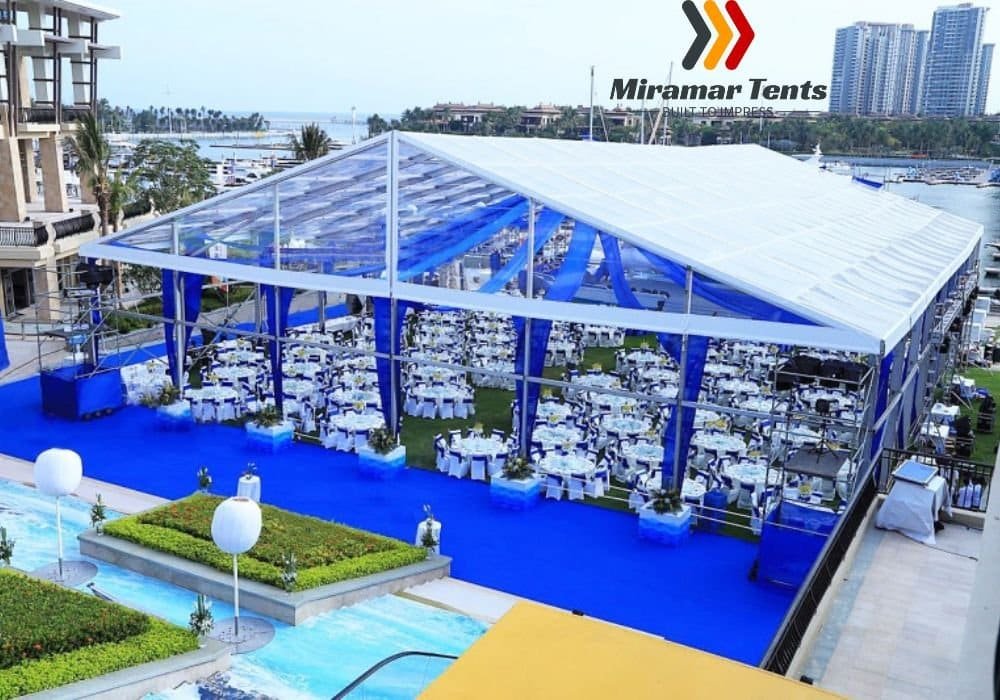 Sentence with replacement: Outdoor event setting with a large All-Occasion Canopy Tent, white chairs and tables, set on a blue carpet near a waterfront, with cityscape in the background.