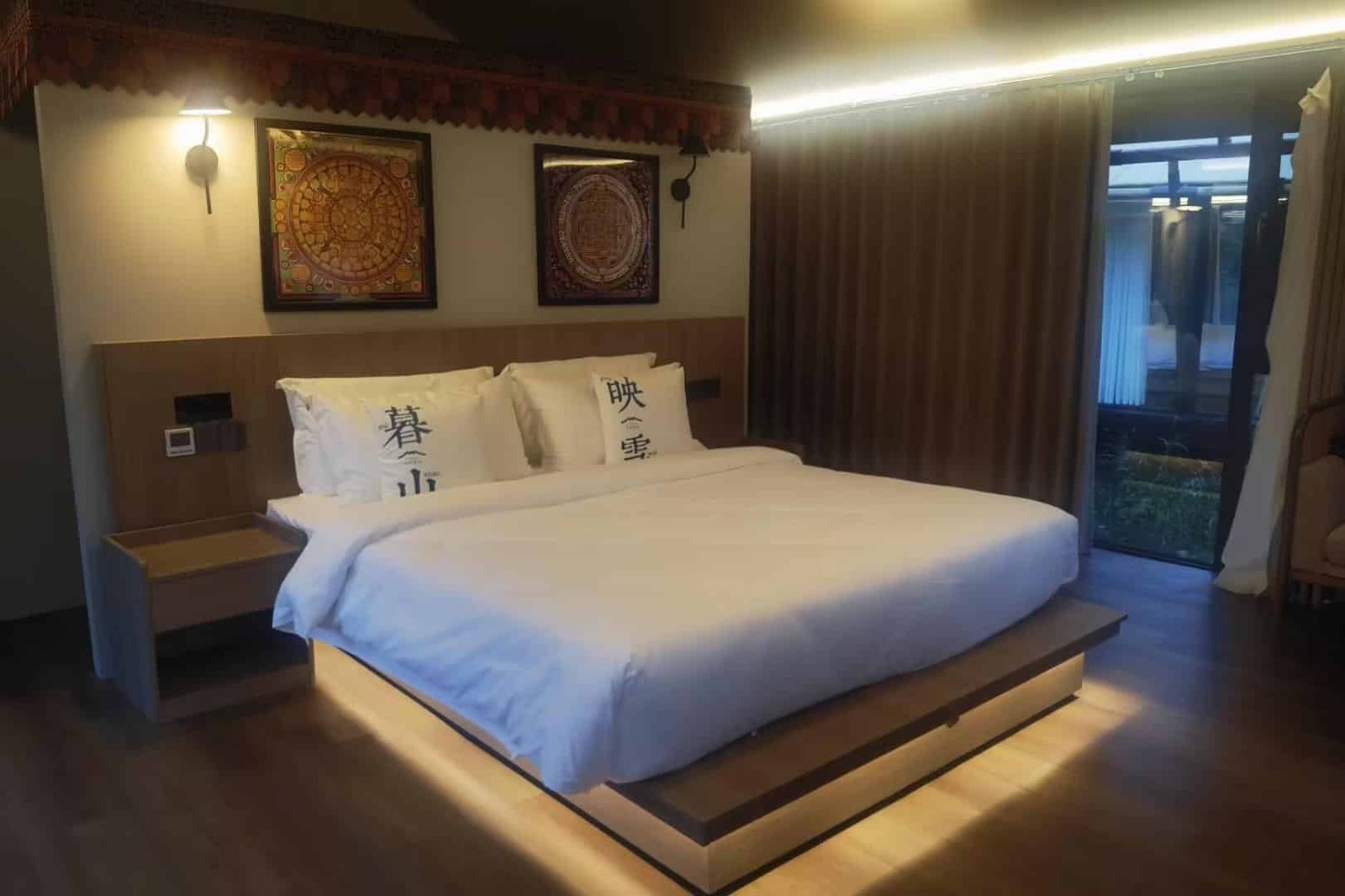 A modern bedroom features a large bed with white linens, two decorative pillows with chinese characters, wall art above the headboard, and floor to ceiling curtains by a window.