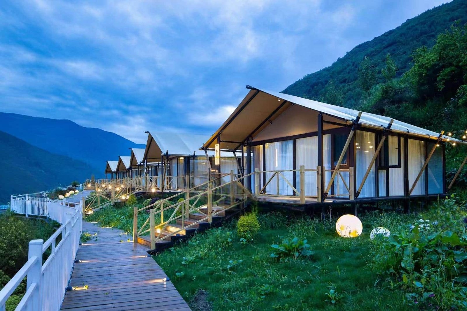 Row of modern glamping cabins with large windows and wooden decks, set on a hillside with a boardwalk and mountain views at dusk.