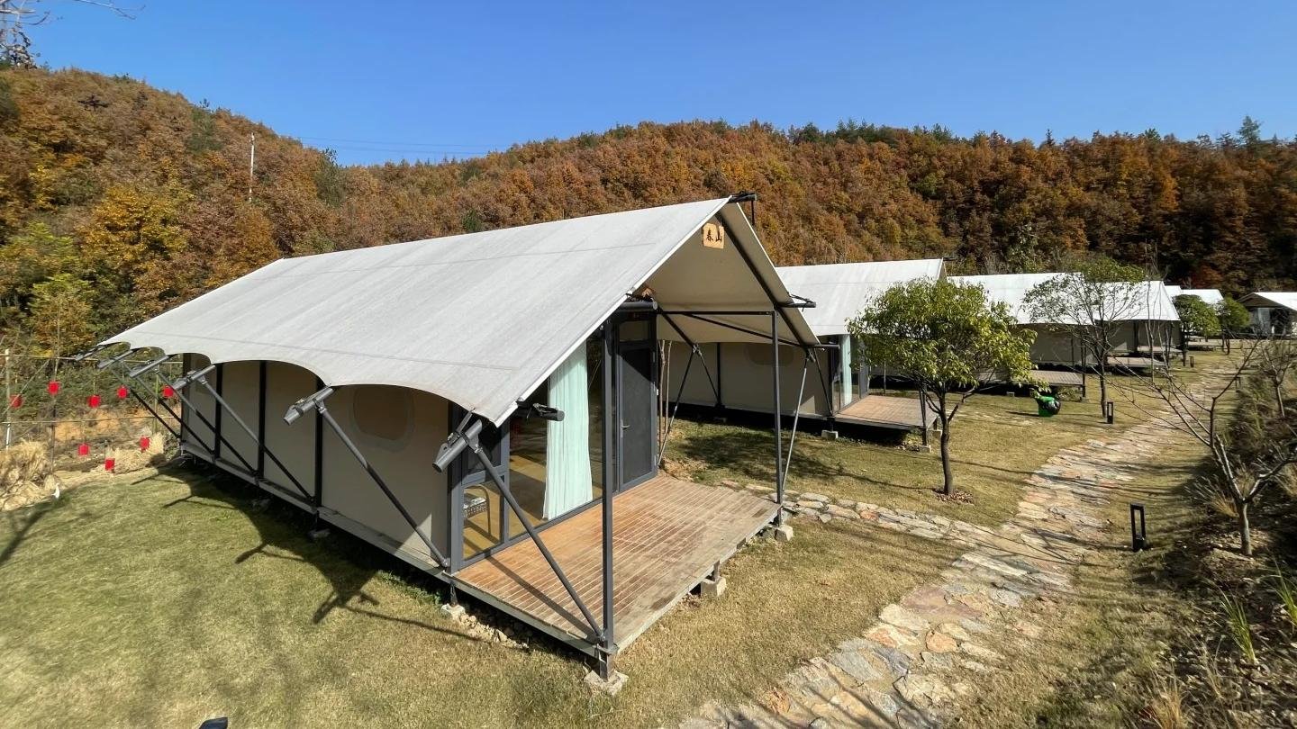 Three canvas glamping tents with wooden decks are set on grassy plots beside a stone path, surrounded by trees with autumn foliage under a clear blue sky.