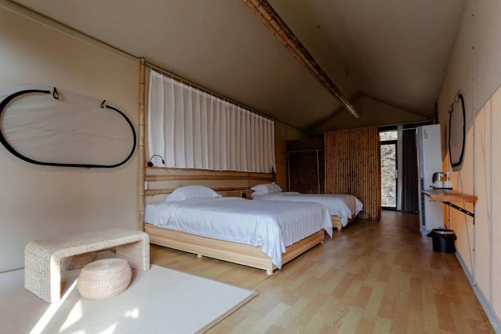 A minimalist room with two beds, light wood flooring, bamboo accents, a small woven table and stool, and a curtained window, with a door and refrigerator in the background.