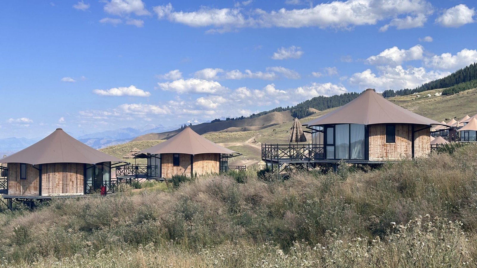 Three round, tent like cabins with brown roofs are situated on a grassy hillside under a partly cloudy sky, surrounded by mountains in the background.