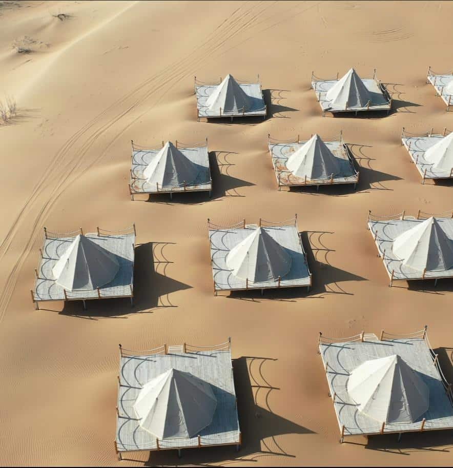 Aerial view of multiple white canvas tents arranged in rows on sandy terrain, casting distinct shadows on the desert ground.