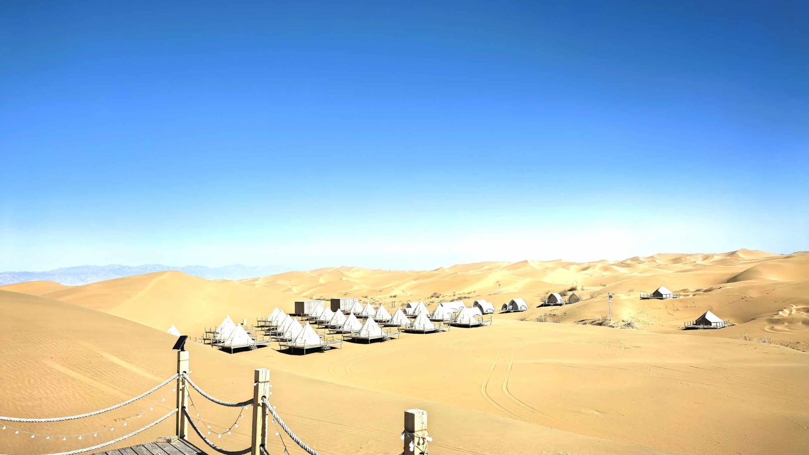 Rows of white tents stand in the middle of a vast desert with rolling sand dunes under a clear blue sky; a rope fence is visible in the foreground.