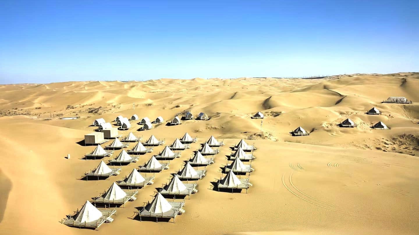 Rows of white tents are set up in a sandy desert landscape under a clear blue sky, surrounded by dunes.