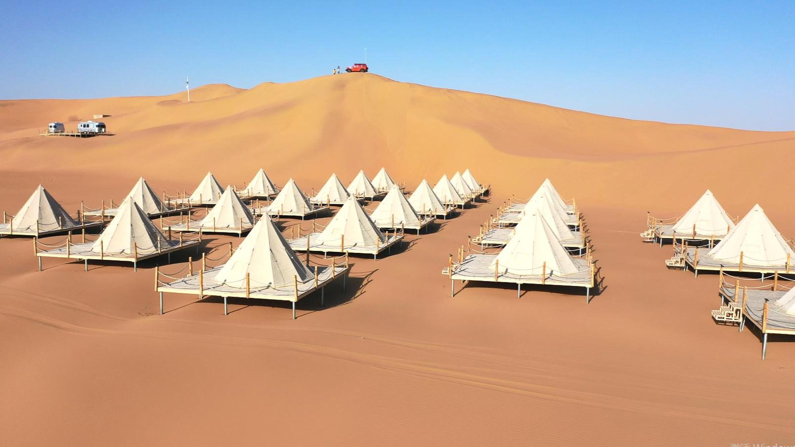Rows of white canvas tents are set up on raised platforms in a sandy desert with dunes and a vehicle visible on a hill in the background under a clear blue sky.