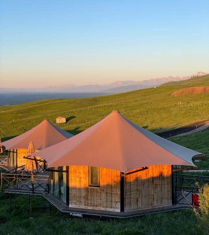 Two modern round wooden huts with conical roofs sit on green grassy hills under a clear sky, with distant mountains visible in the background.