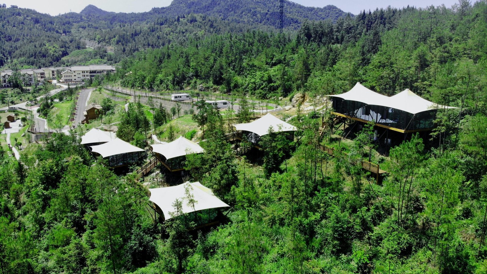 A cluster of white roofed structures is surrounded by dense green forest on a hillside, with mountains and additional buildings visible in the background.