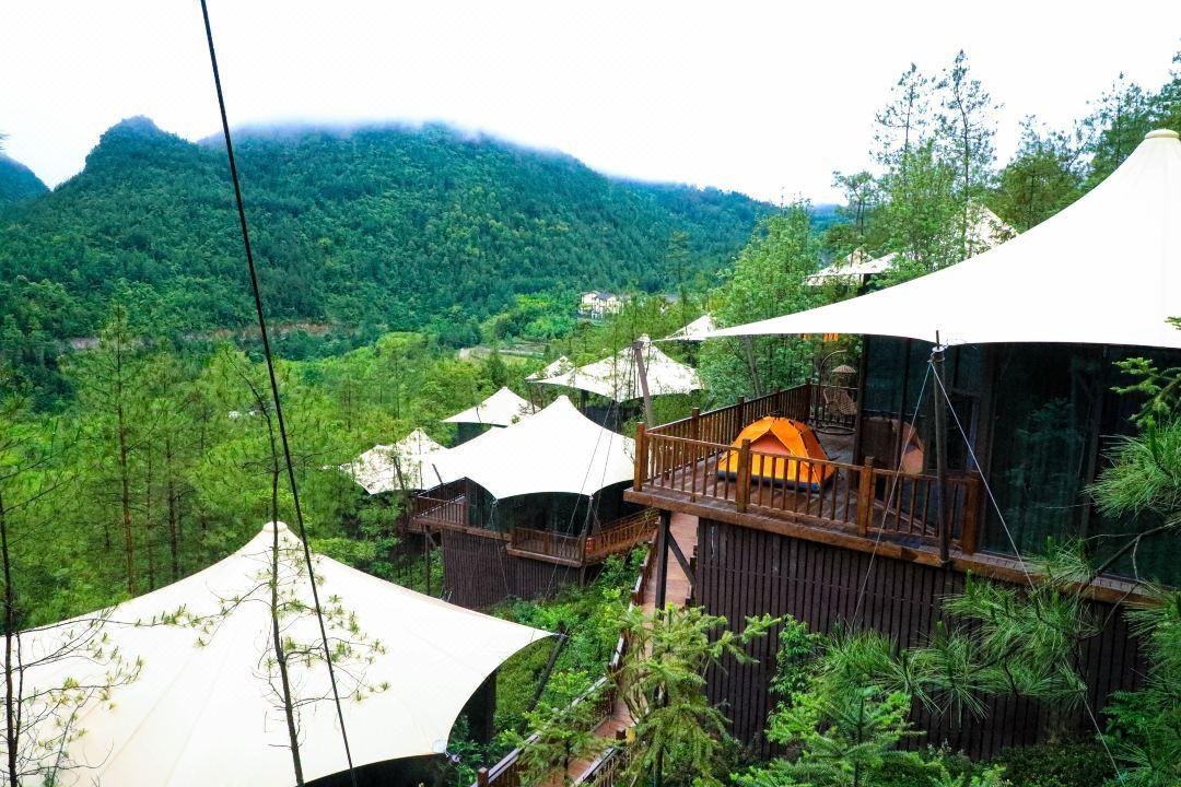 Elevated cabins with white tent roofs are nestled among trees on a green hillside, with mountains visible in the background.