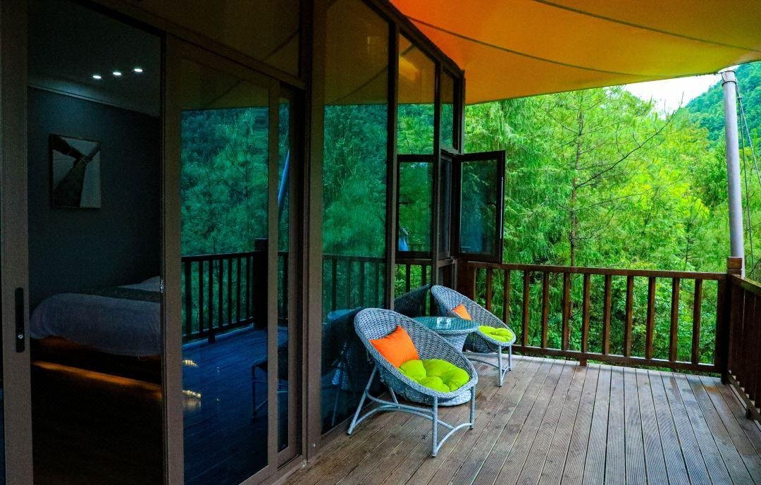 A wooden balcony with two wicker chairs and orange cushions overlooks green trees; a glass door leads to a bedroom inside.