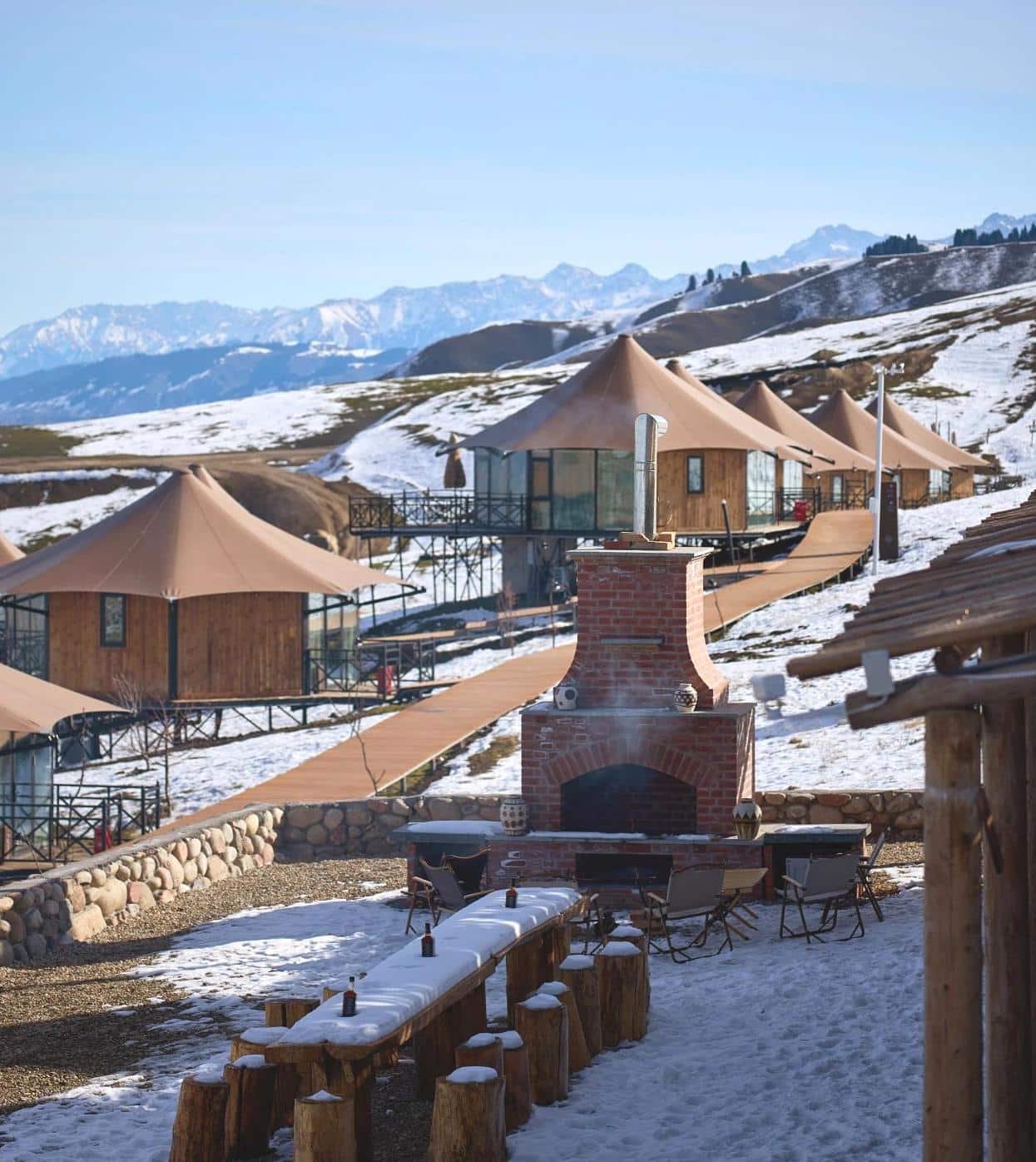 A row of wooden cabins with canvas roofs on a snowy mountain slope, with outdoor seating and a brick fireplace in the foreground, and mountains in the background.