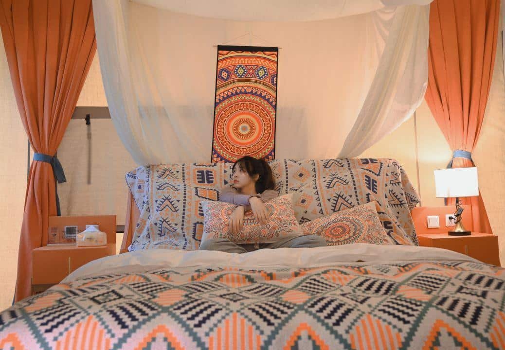 A person sits on a bed with patterned bedding in a room decorated with warm colors, geometric designs, and soft lighting.