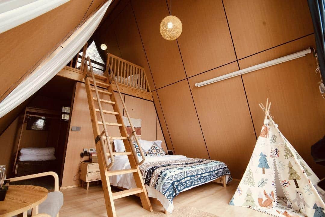 A cozy a frame cabin interior featuring a loft bed accessed by a wooden ladder, a double bed, a patterned teepee, light wood furniture, and a hanging lamp.