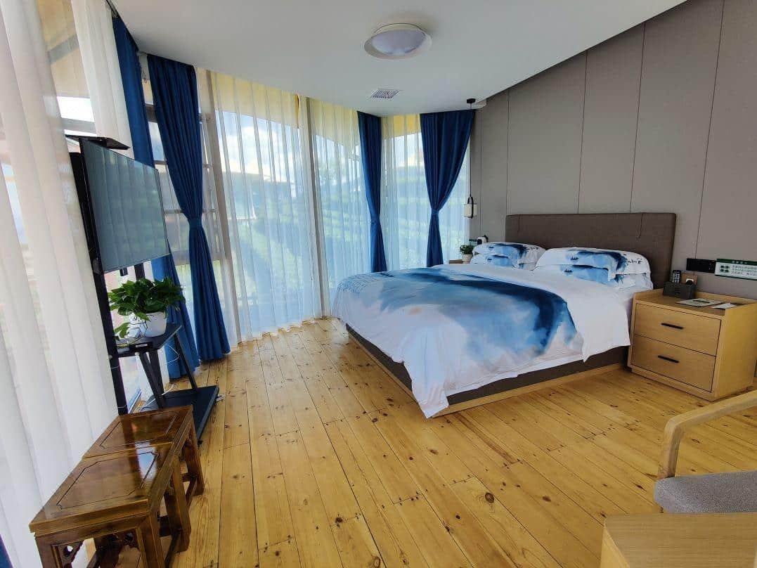 A modern bedroom with large windows, wooden floor, a double bed with blue and white bedding, a tv, and a small nightstand. blue curtains frame the windows.
