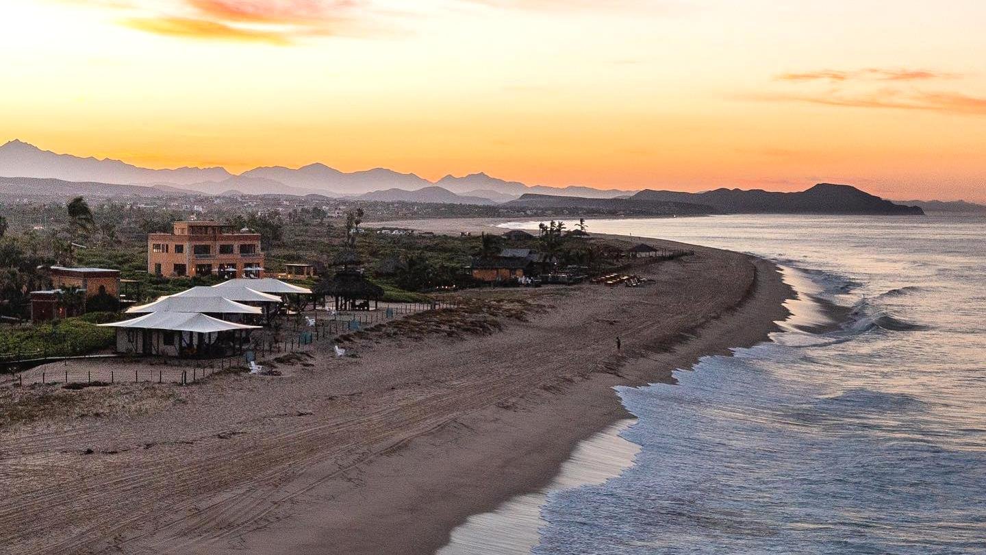 A coastal scene at sunset shows gentle waves, a sandy beach, several buildings, and distant mountains under an orange sky.