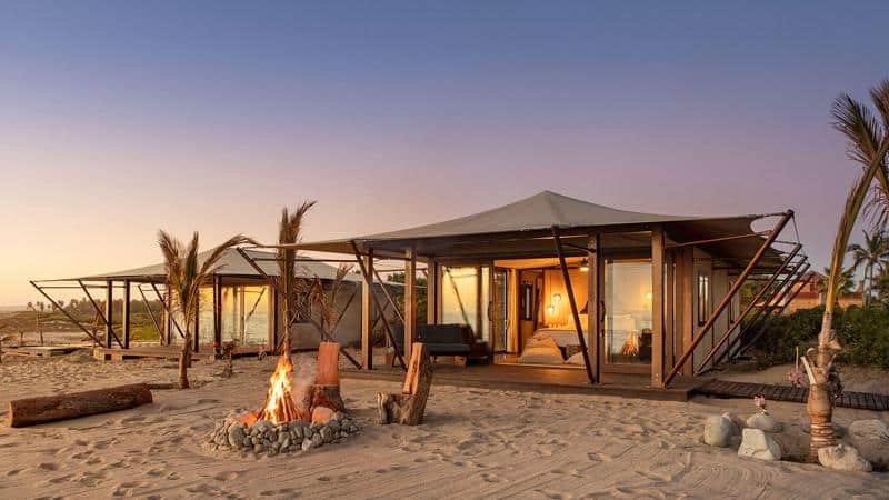 Modern beachside cabanas with canvas roofs and glass walls sit on sandy ground, with a lit campfire and scattered logs in the foreground at sunset.