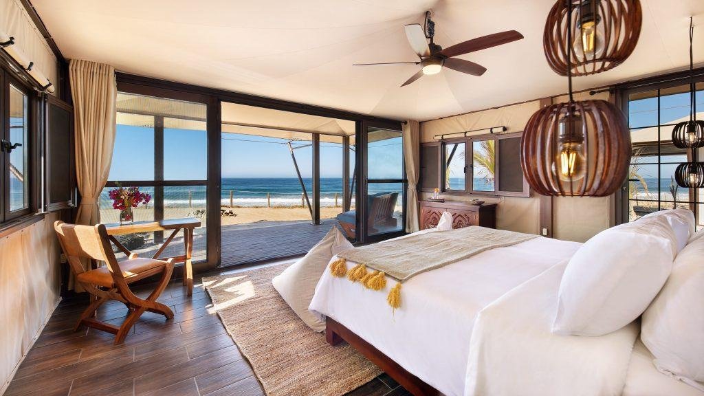 A bright bedroom with a large bed, wooden furniture, hanging lights, and glass doors opening to an ocean view and deck.