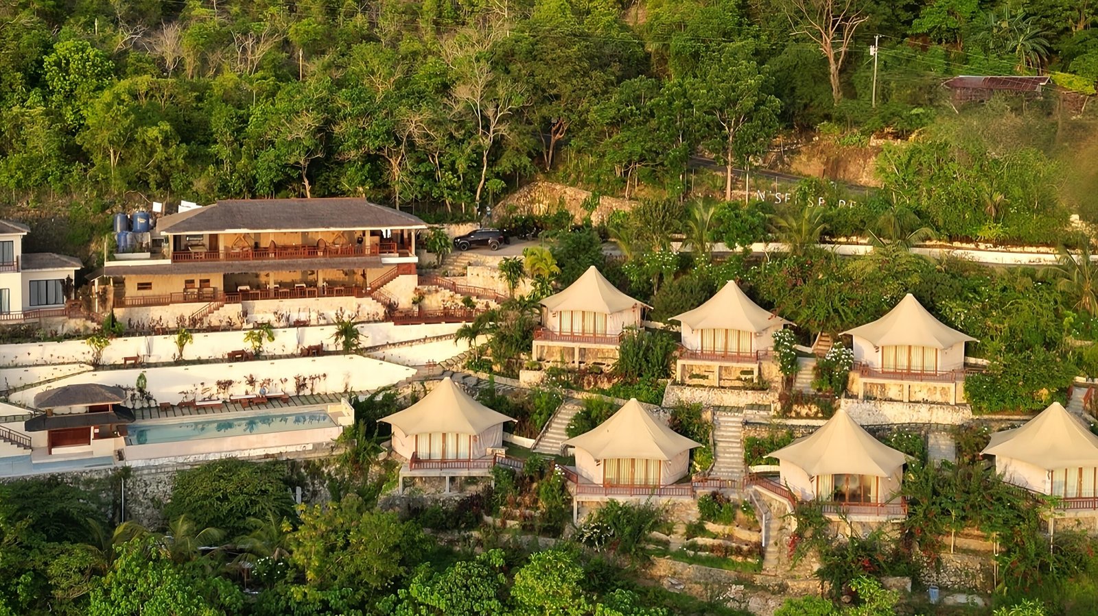 A hillside resort with several tent shaped villas, a swimming pool, and a main building, surrounded by lush green trees and vegetation.