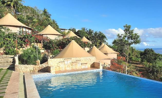 Circular tented cottages and stone walls sit beside an outdoor infinity pool, surrounded by lush greenery with a view of the ocean under a clear blue sky.