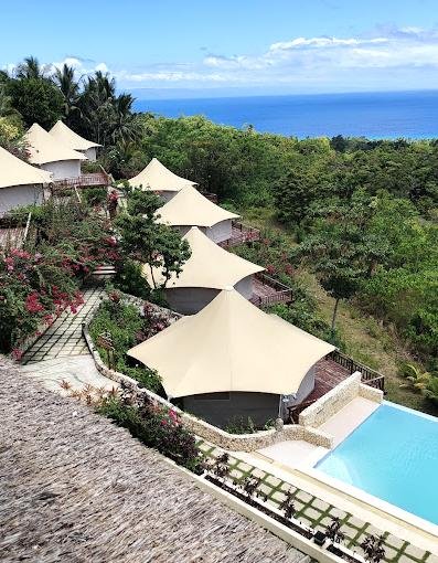 A row of tent like villas on a hillside overlooks a pool and lush greenery, with the ocean visible in the background under a partly cloudy sky.