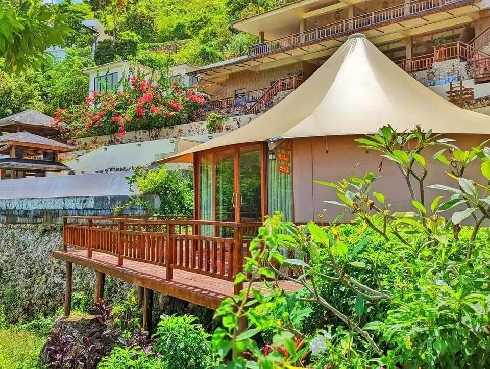 A modern bungalow with a large canopy roof and wooden deck is surrounded by lush greenery and flowering plants on a hillside. multiple terraces with railings are visible above.