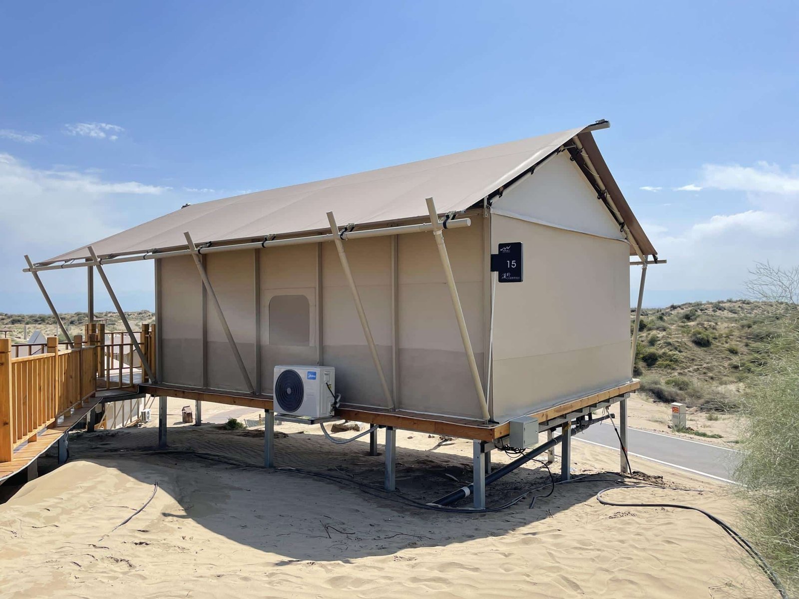 A beige, canvas sided cabin on stilts stands on sandy ground with an air conditioning unit attached, near a wooden walkway, in a dry, open landscape.