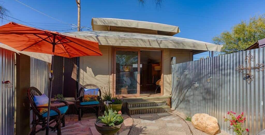 Small patio with two wicker chairs and a table, red umbrella, potted plants, and corrugated metal fencing, leading to a sliding glass door under a canvas awning.