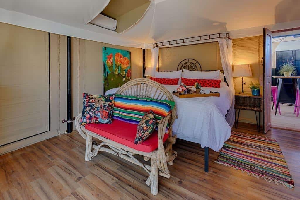 Cozy bedroom with a canopy bed, colorful pillows and bench, wooden floors, a bedside lamp, vibrant rug, and flower painting on the wall.