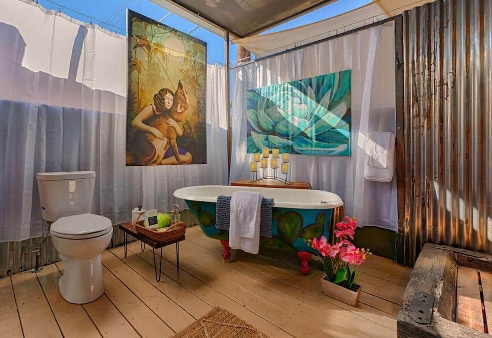 An eclectic bathroom with a freestanding bathtub, toilet, wooden floor, metal walls, colorful artwork, flowers, and candles, with natural light coming through the ceiling.