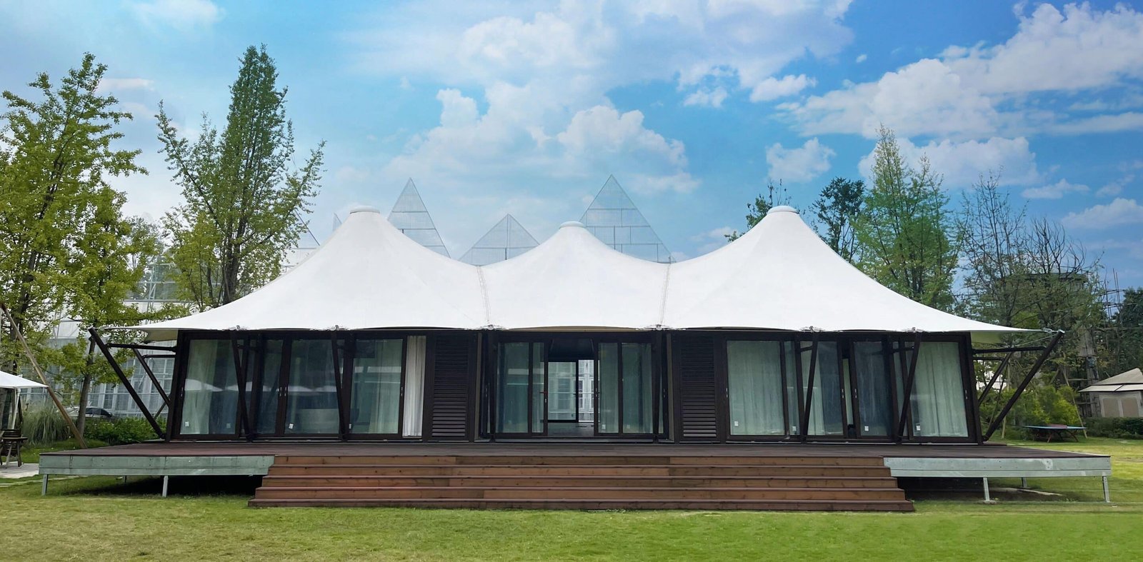 A modern tent like building with a white canopy roof, dark frame, glass walls, and wooden steps, set on a grassy area with trees and a partly cloudy sky in the background.