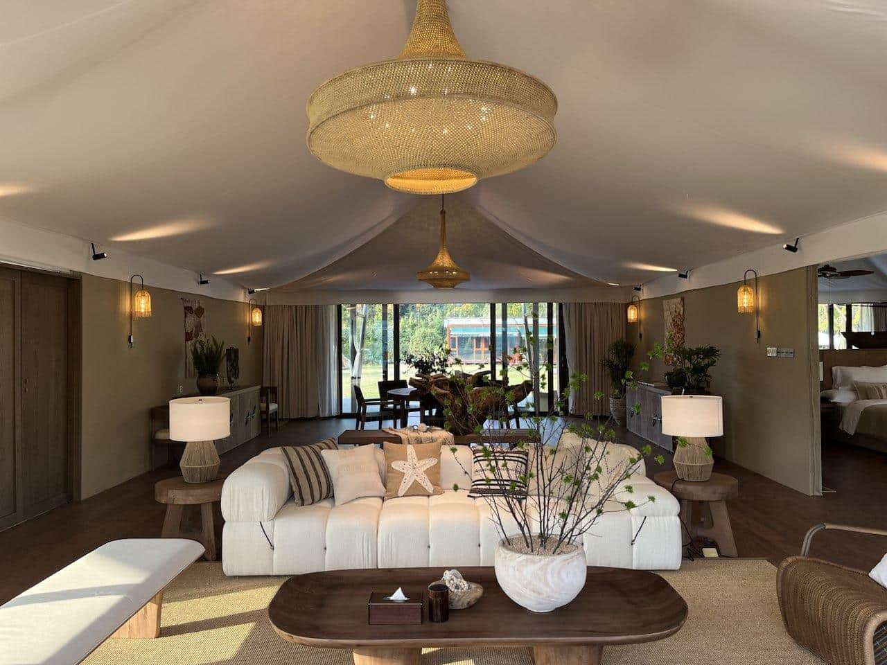 Spacious living room with beige sofas, wooden tables, plants, and large pendant lights under a tent like ceiling, with sliding glass doors opening to a garden view.
