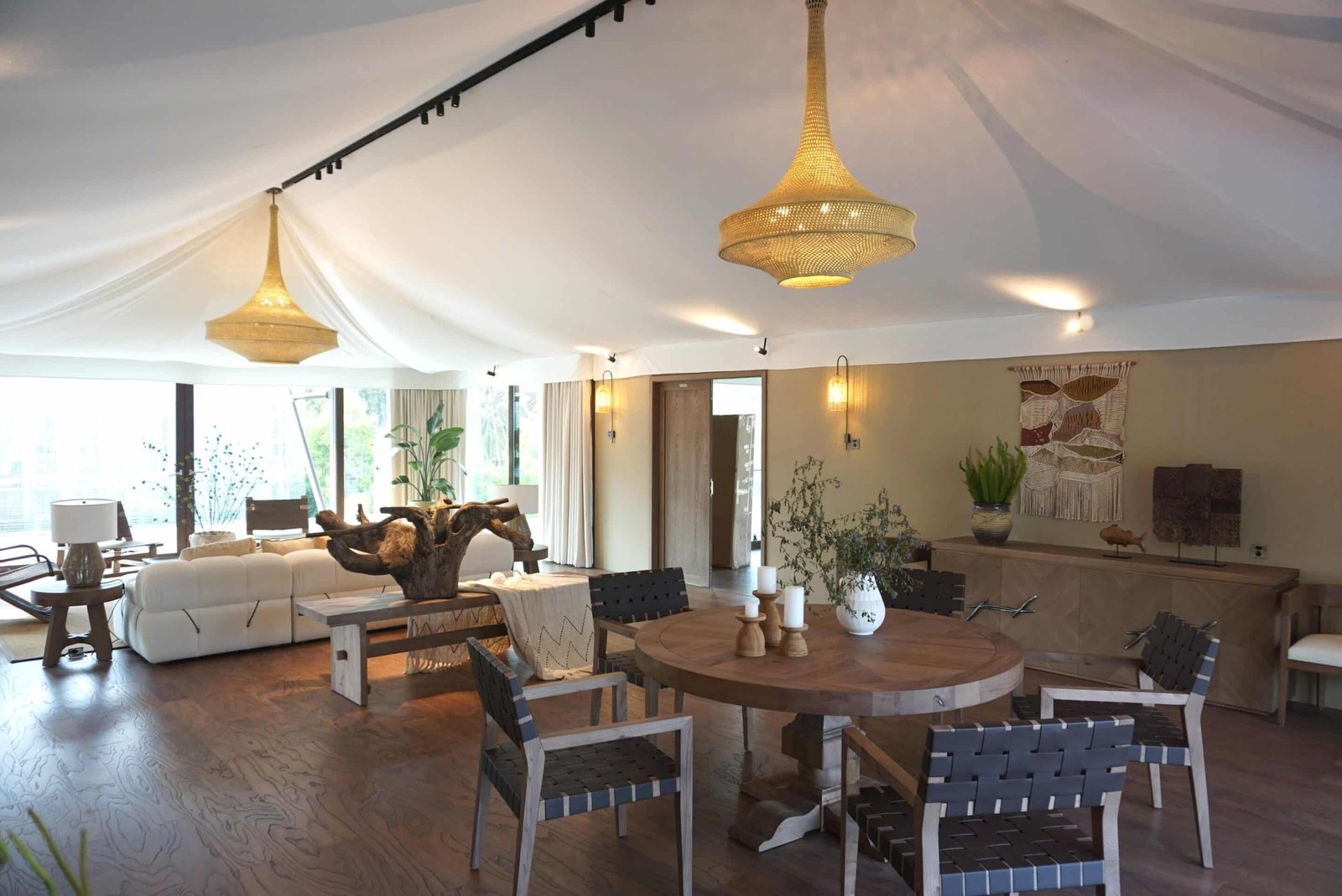 Spacious living room with wooden furniture, round dining table, white sofas, large windows, decorative plants, and two woven pendant lights under a draped ceiling.