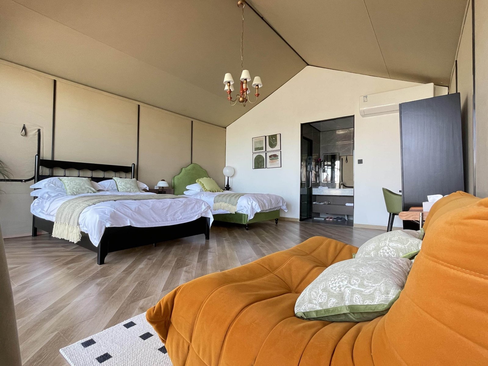 A spacious bedroom features two beds, a mustard colored sofa, a desk, a wardrobe, light wood flooring, and an ensuite bathroom visible in the background.