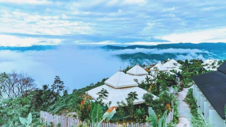 A row of white tents and cottages surrounded by greenery on a hillside, overlooking a misty valley and distant mountains under a cloudy sky.