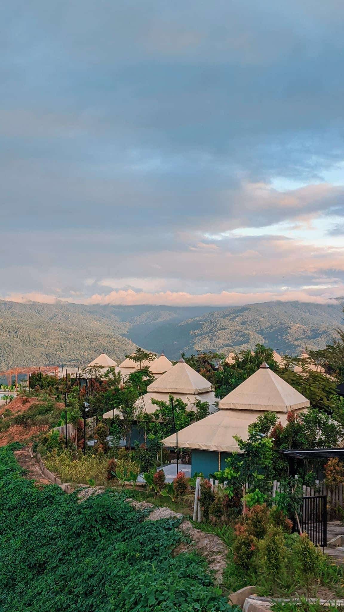 A row of white tents sits among greenery on a hillside, with mountains and a partly cloudy sky in the background.