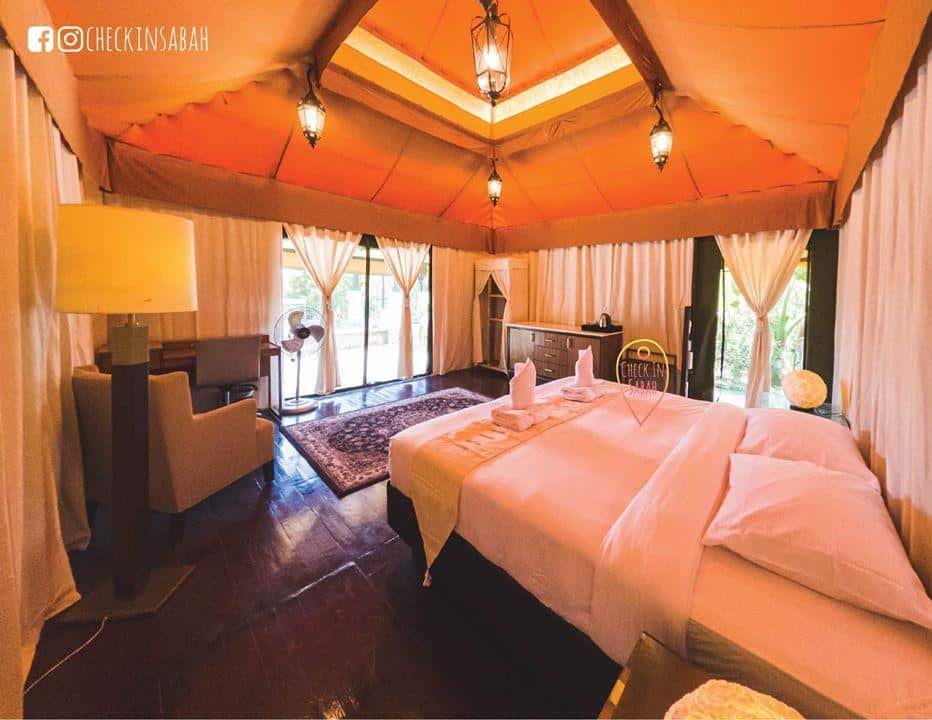 A spacious hotel room with a large bed, armchair, desk, fan, rug, and large windows with sheer curtains letting in natural light.