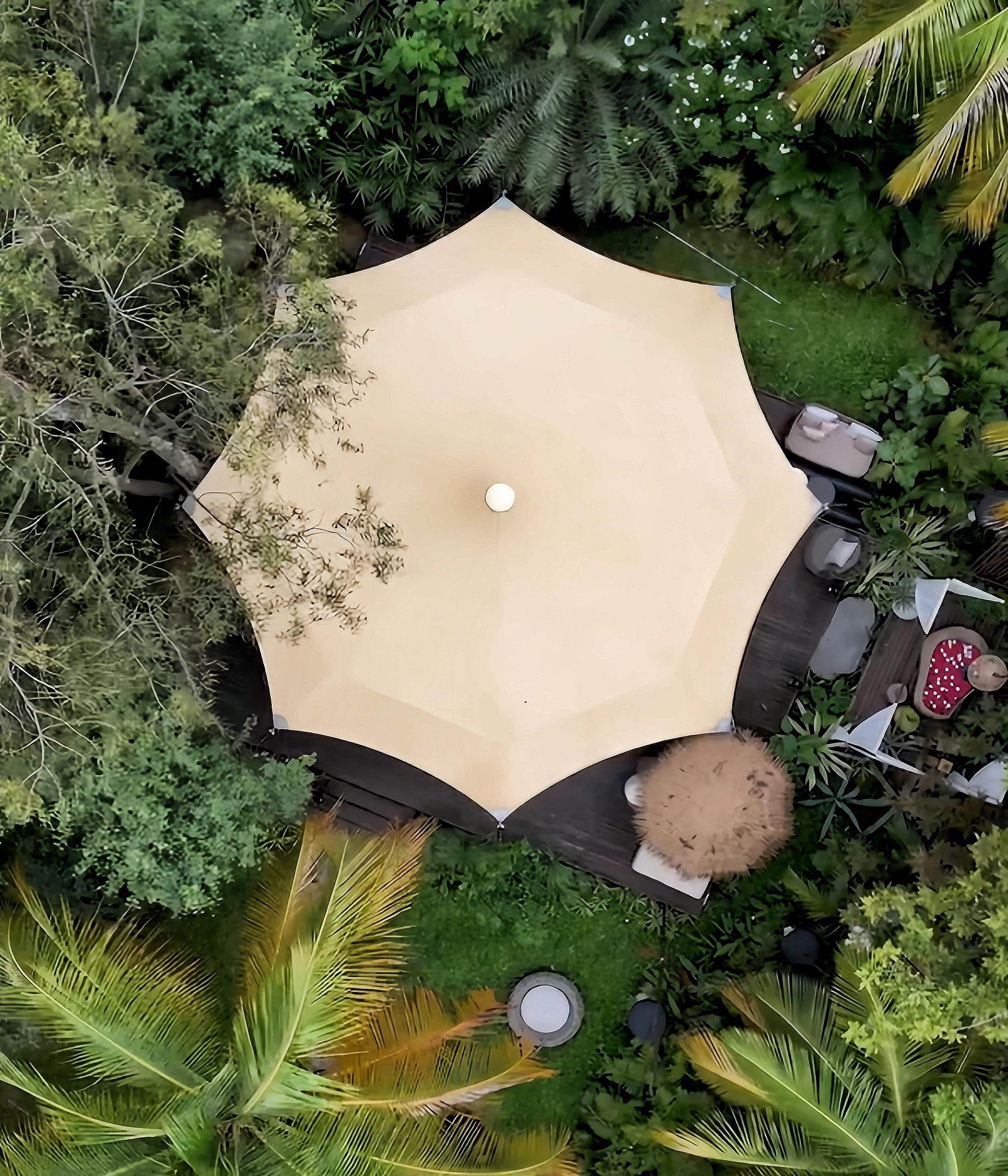 A large beige patio umbrella shades an outdoor seating area on a wooden deck, surrounded by lush green trees and plants, viewed from above.