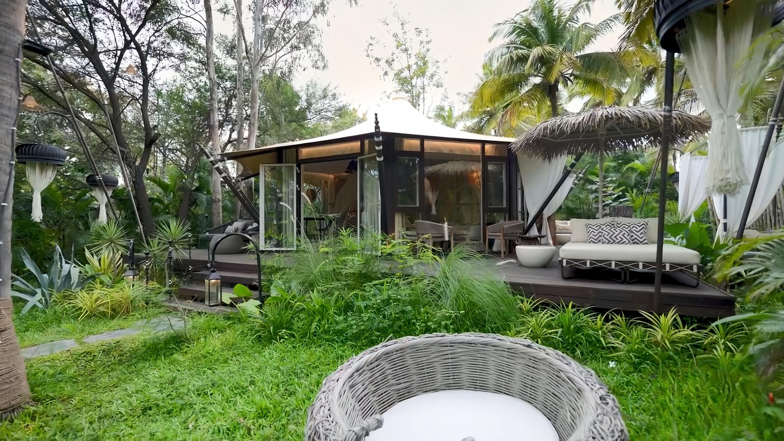 Modern glass cabin with a patio surrounded by lush greenery, palm trees, outdoor furniture, and a wicker chair in the foreground.