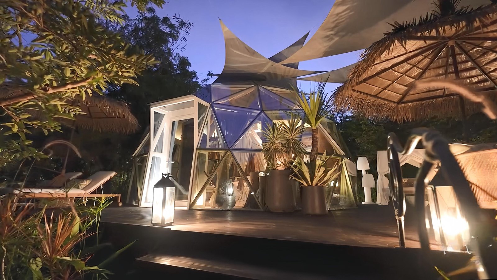 A modern glass geodesic dome with a wooden deck, surrounded by tropical plants, lounge chairs, lanterns, and thatched umbrellas at dusk.