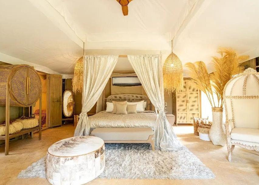 A bedroom with a canopy bed, white curtains, and neutral toned decor, including a round ottoman, shag rug, wicker accents, and tall dried plants.