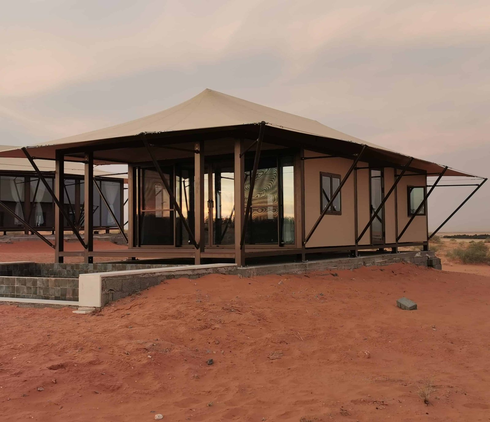 A modern tent style cabin with glass walls and a beige roof stands on a raised stone platform in a desert landscape at sunset.