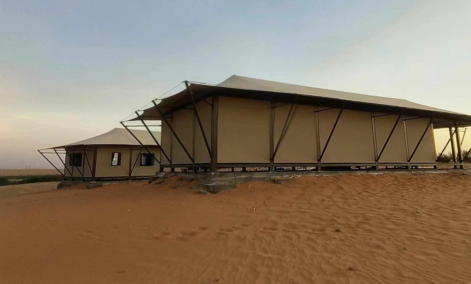 Two beige canvas tents with metal frames sit on a sandy desert terrain at sunset, with a clear sky in the background.