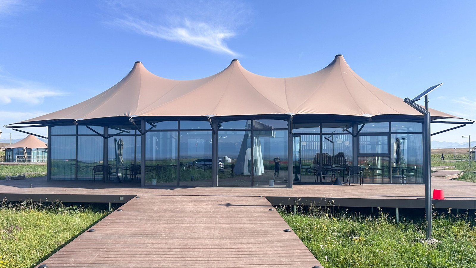 A modern tent like glass building with a beige canopy roof sits on a wooden deck in a grassy outdoor setting under a blue sky.