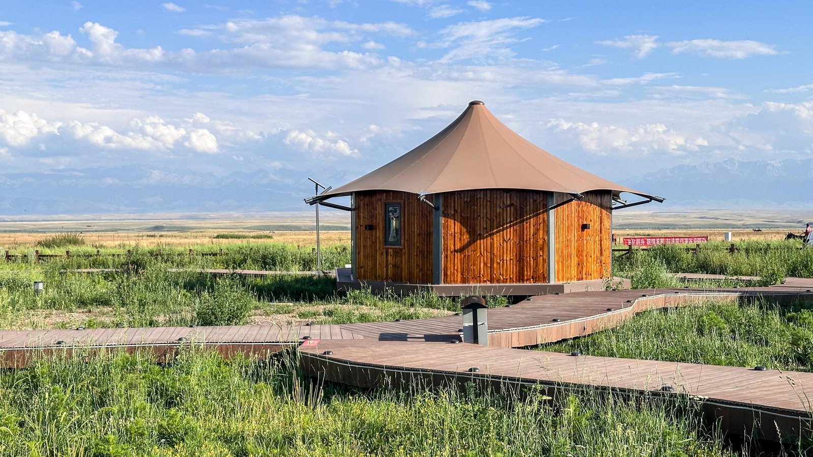 A round wooden cabin with a conical roof sits on a wooden boardwalk surrounded by green grass, with mountains and a cloudy sky in the background.