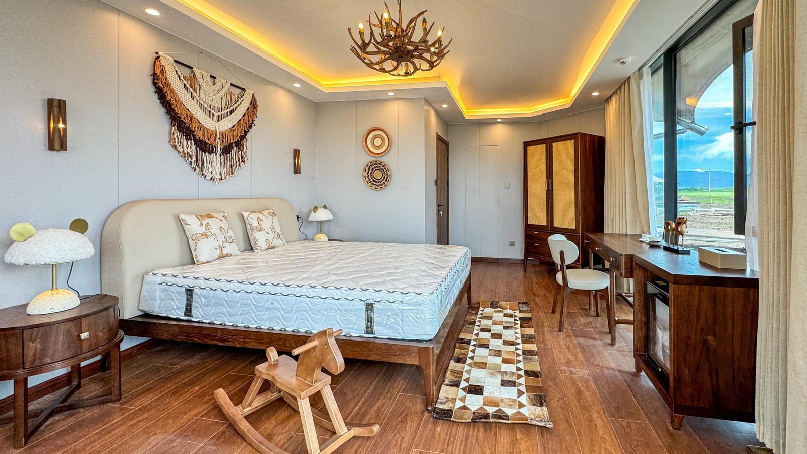 A modern bedroom with a large bed, wooden furniture, a rocking horse, wall decor, and large windows with a view of the outdoors. warm lighting accents the ceiling.
