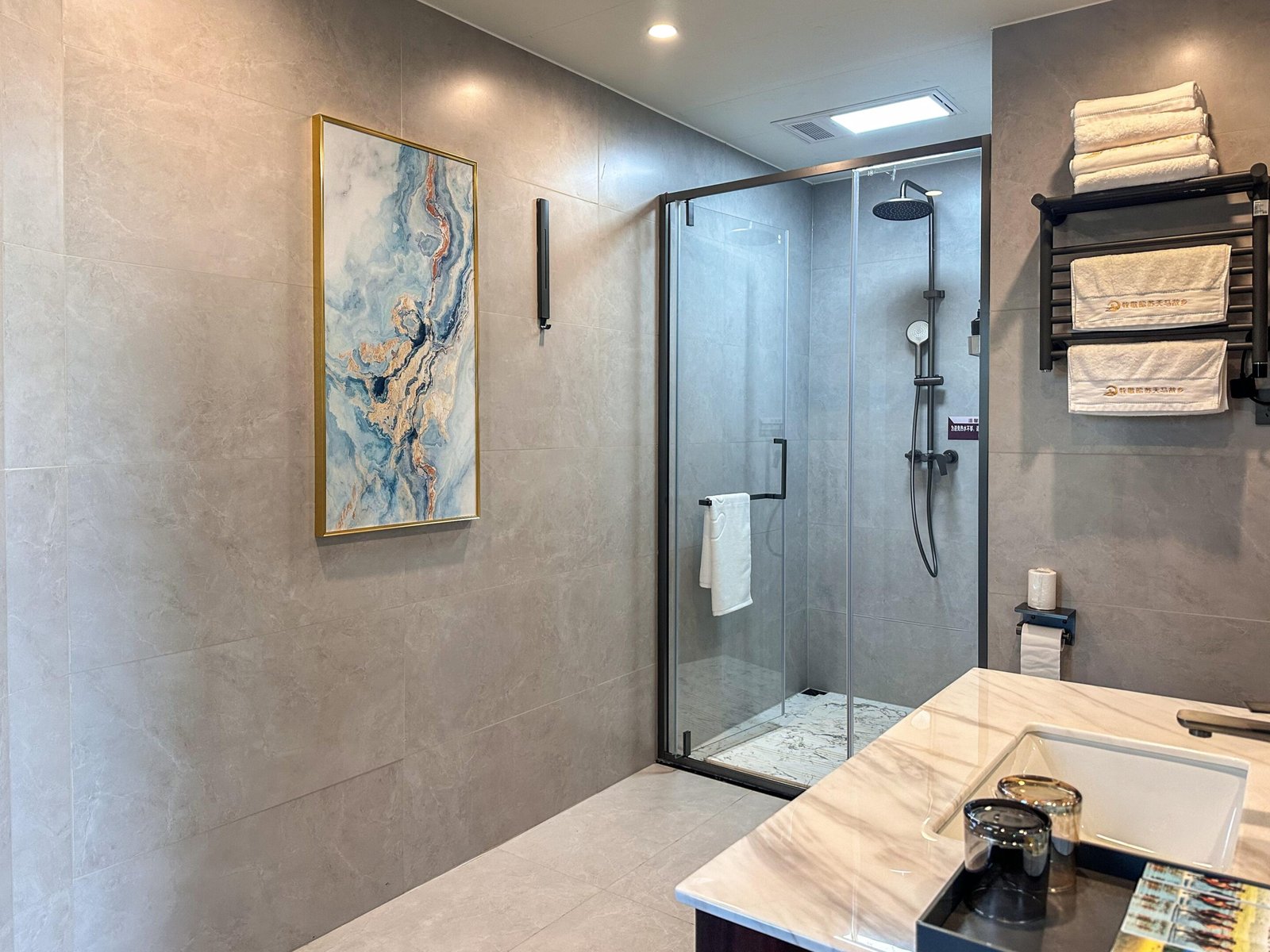 Modern bathroom with gray tiled walls, a glass shower enclosure, a wall mounted towel rack with folded towels, abstract wall art, and a white marble countertop with a sink.