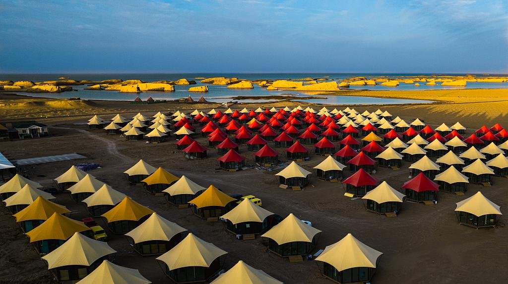 Rows of red, yellow, and beige tents set up on a sandy landscape near water, with rock formations and blue sky in the background.