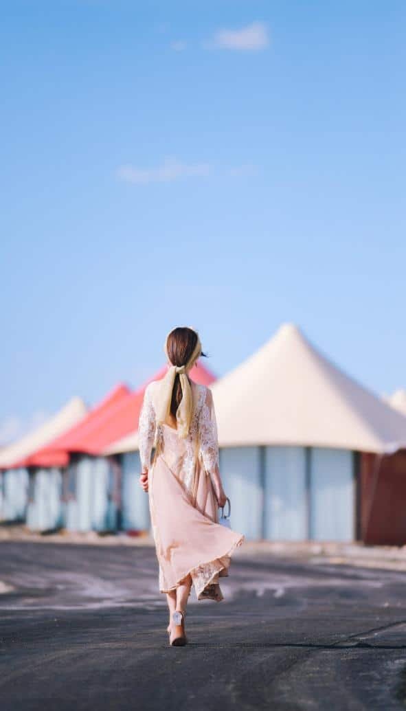 A woman in a light dress walks away on a paved path toward tent like structures under a clear blue sky.