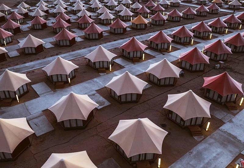Aerial view of rows of large, dome shaped tents with beige and pink tops arranged uniformly on a sandy ground with walkways between them.