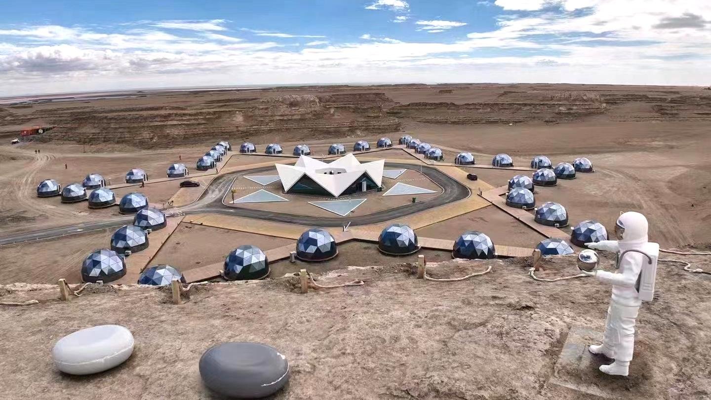 A person in a white spacesuit stands on a rocky ledge overlooking a futuristic dome shaped camp set up in a barren desert landscape.